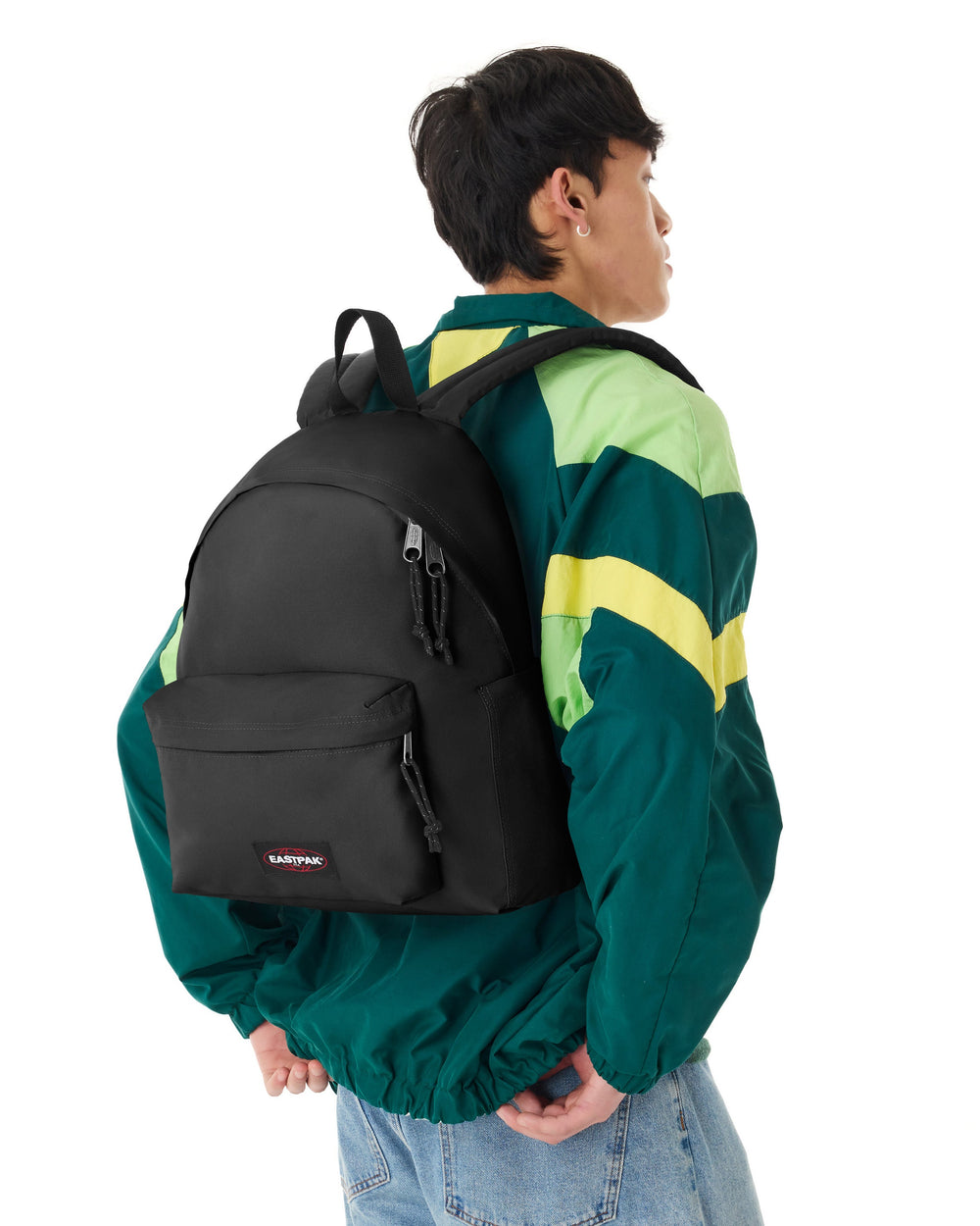 Eastpak mochila Day Pak’r negra a la espalda, con una chaqueta verde y amarilla y unos vaqueros azul claro.