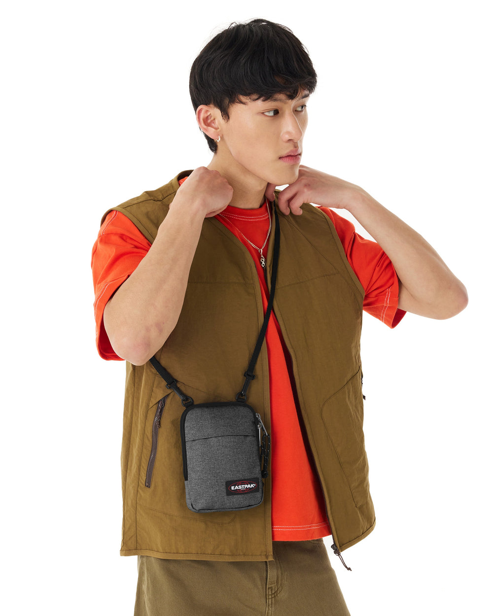 Eastpak TH grey crossbody bag worn over an orange T-shirt and a khaki vest.