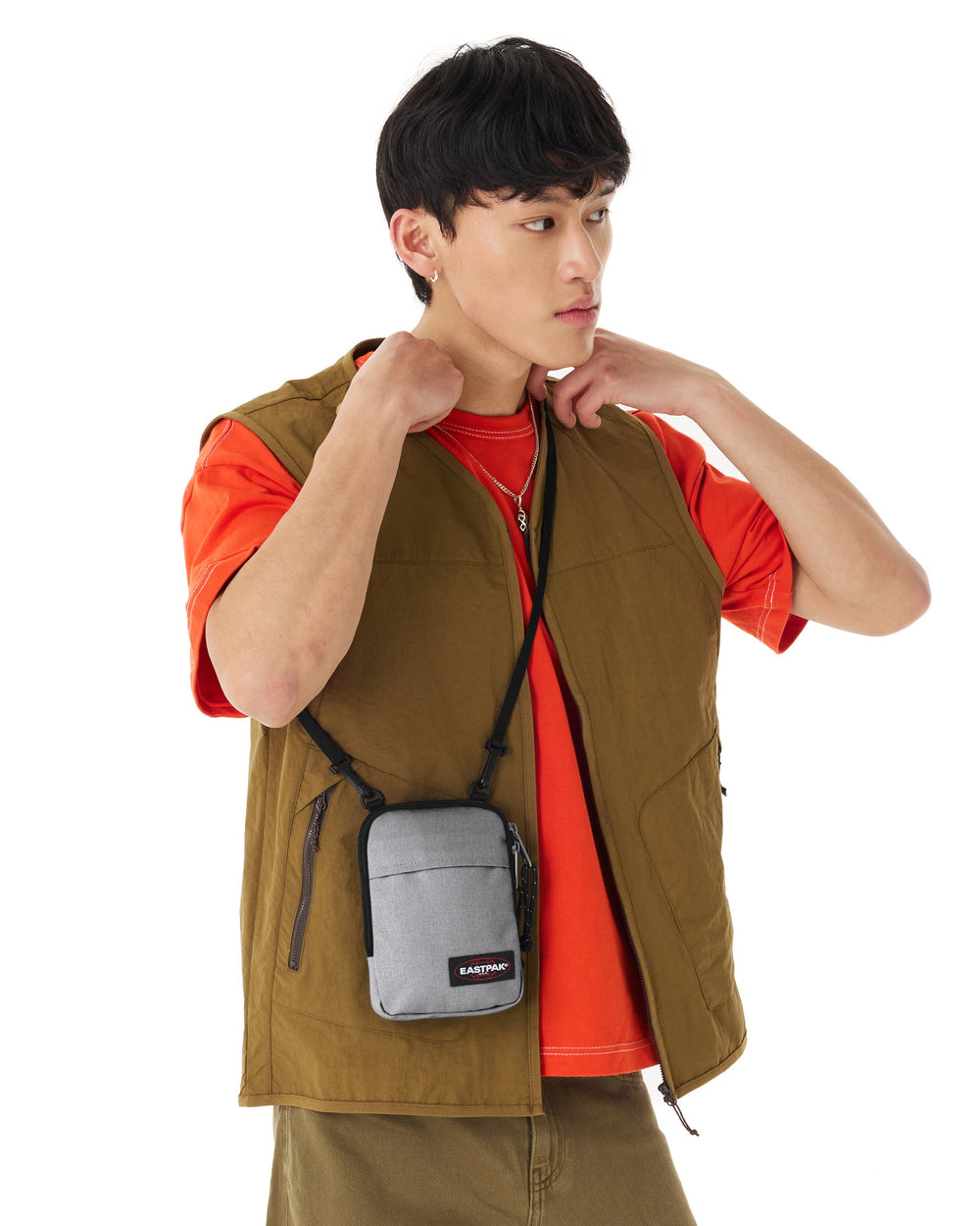 Eastpak The One grey crossbody bag, worn with an orange T-shirt and a khaki vest.