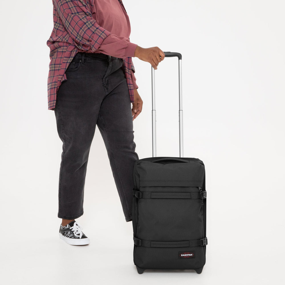 Eastpak čierna taška Transit’R S, držaná za teleskopickú rukoväť, s ružovou kockovanou košeľou a čiernymi nohavicami.