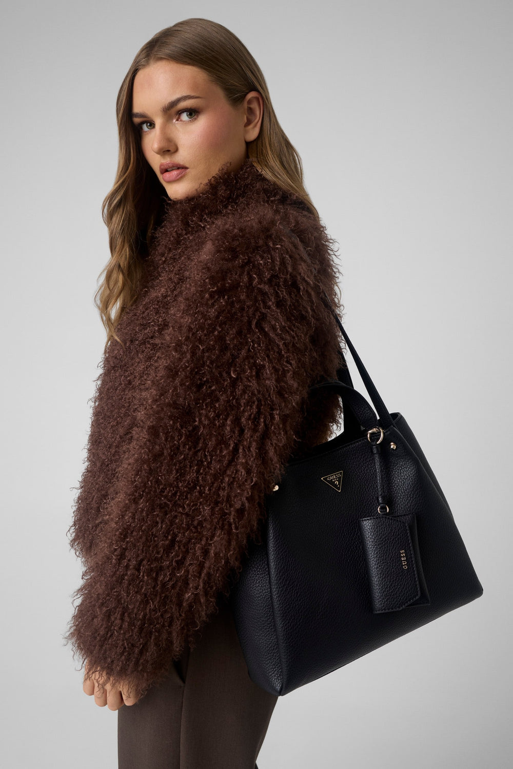 Guess zwarte Lorella-shopper over de schouder, met een bruine broek en ruimvallend jasje.