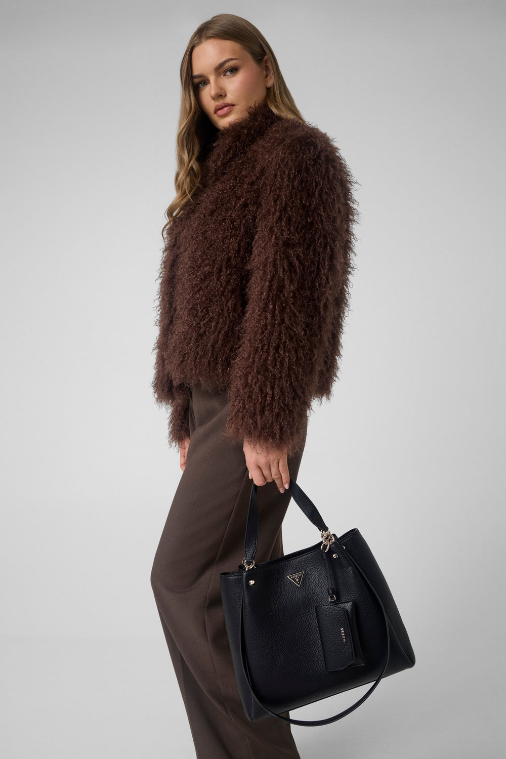 Guess zwarte Lorella-shopper in de hand gedragen, met een bruine jas en bijpassende broek.