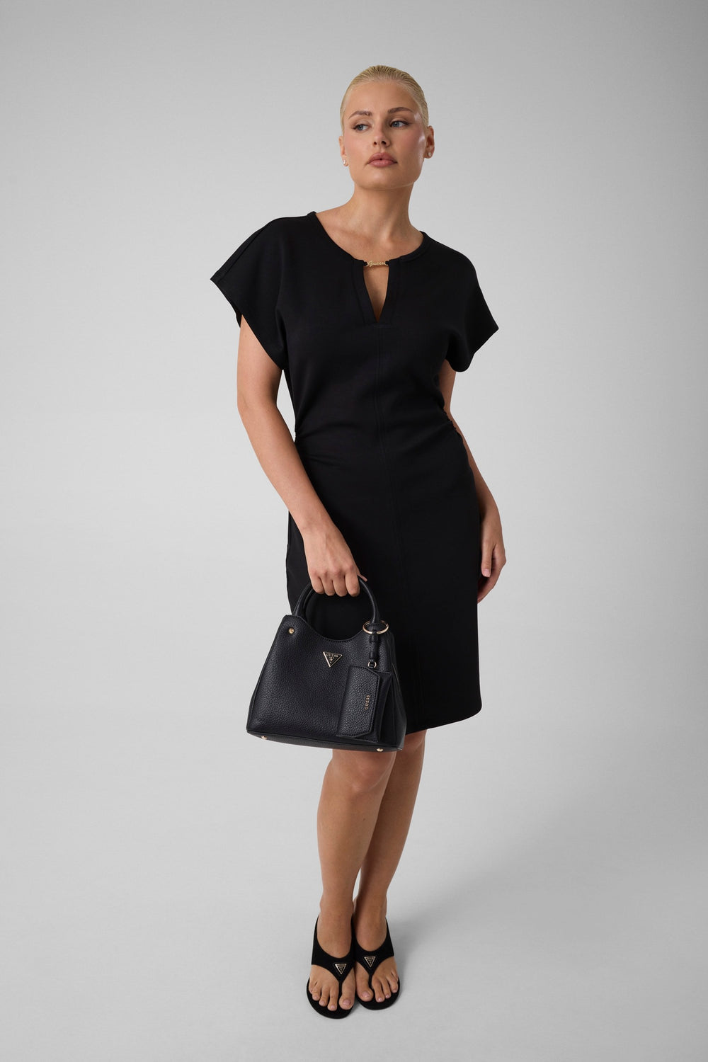 Guess schwaarze Shopper Lorella Petit an der Hand, mat engem schwaarze Kleed a schwaarze Sandalen.