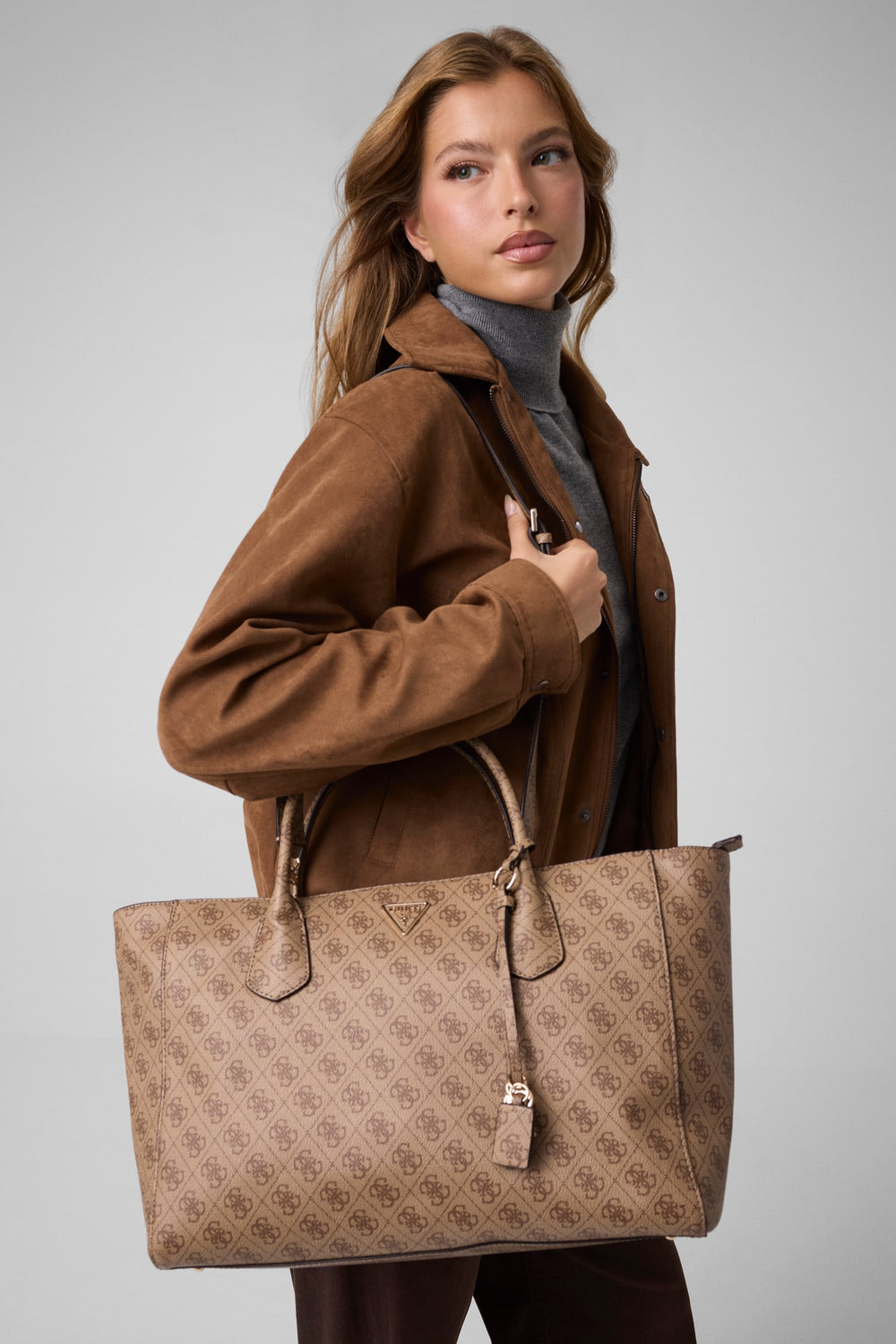 Guess tote Marissa Monogramme beige tenuta a mano con una giacca marrone e un dolcevita grigio.