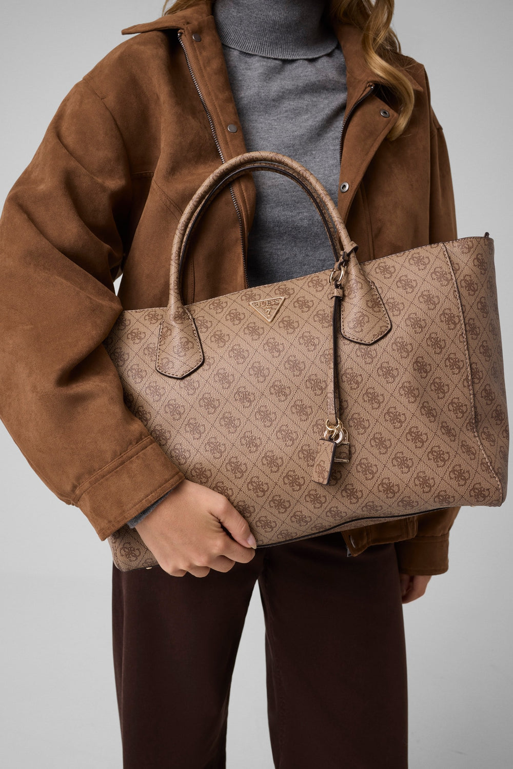 Guess shopper Marissa Monogramme beige tenuta a mano, con giacca marrone, dolcevita grigio e pantaloni marroni.