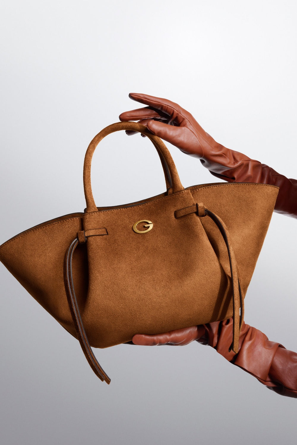 Guess Allegra tote bag in brown, held by hand against a light gray background.