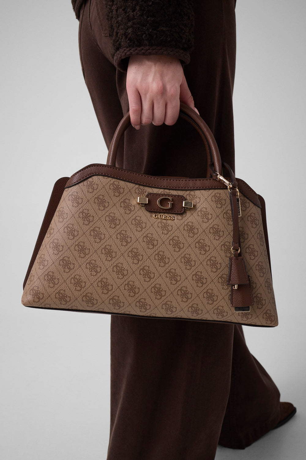 Guess beige handtas Lindsey, in de hand gehouden bij een bruine broek en een donker jasje.