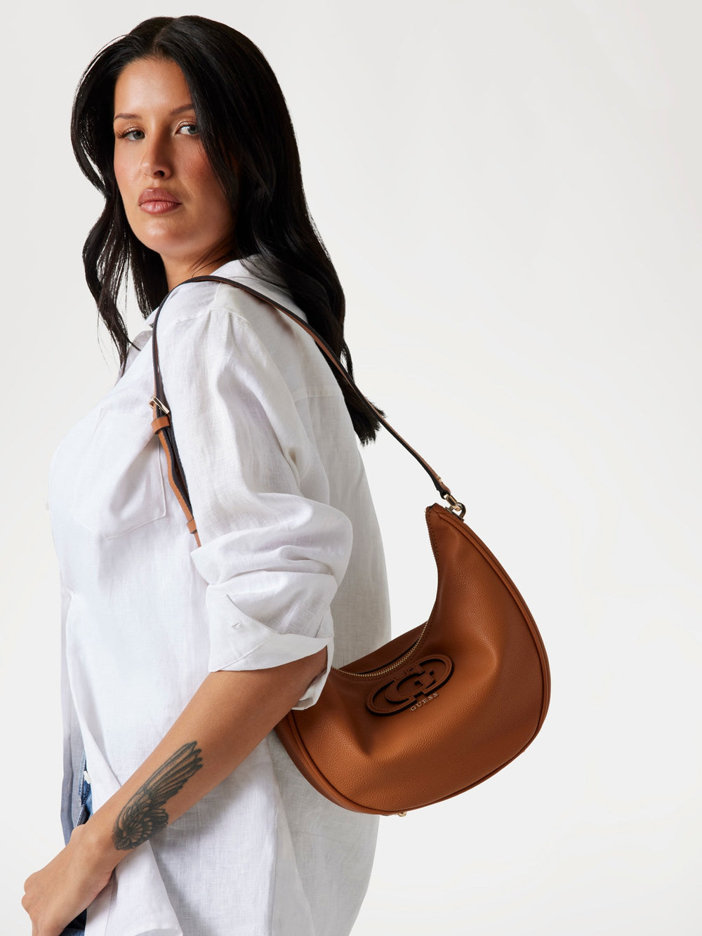 Guess sac seau Silvye Bucket marron porté à l’épaule avec une chemise blanche et un jean bleu.
