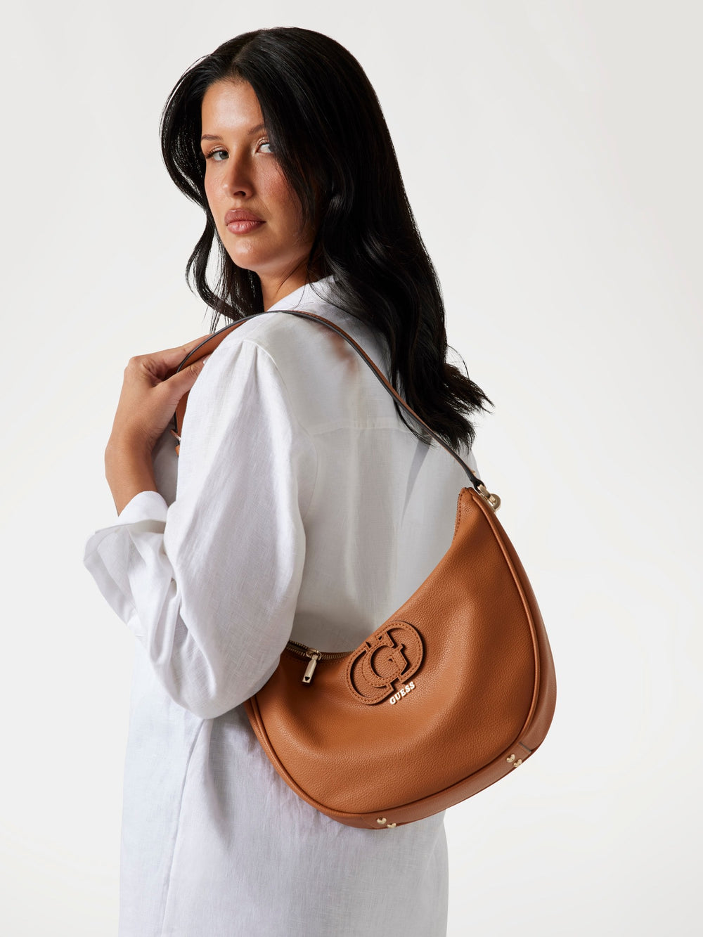 Guess sac seau Silvye Bucket marron porté à l’épaule avec une chemise blanche et un pantalon clair.
