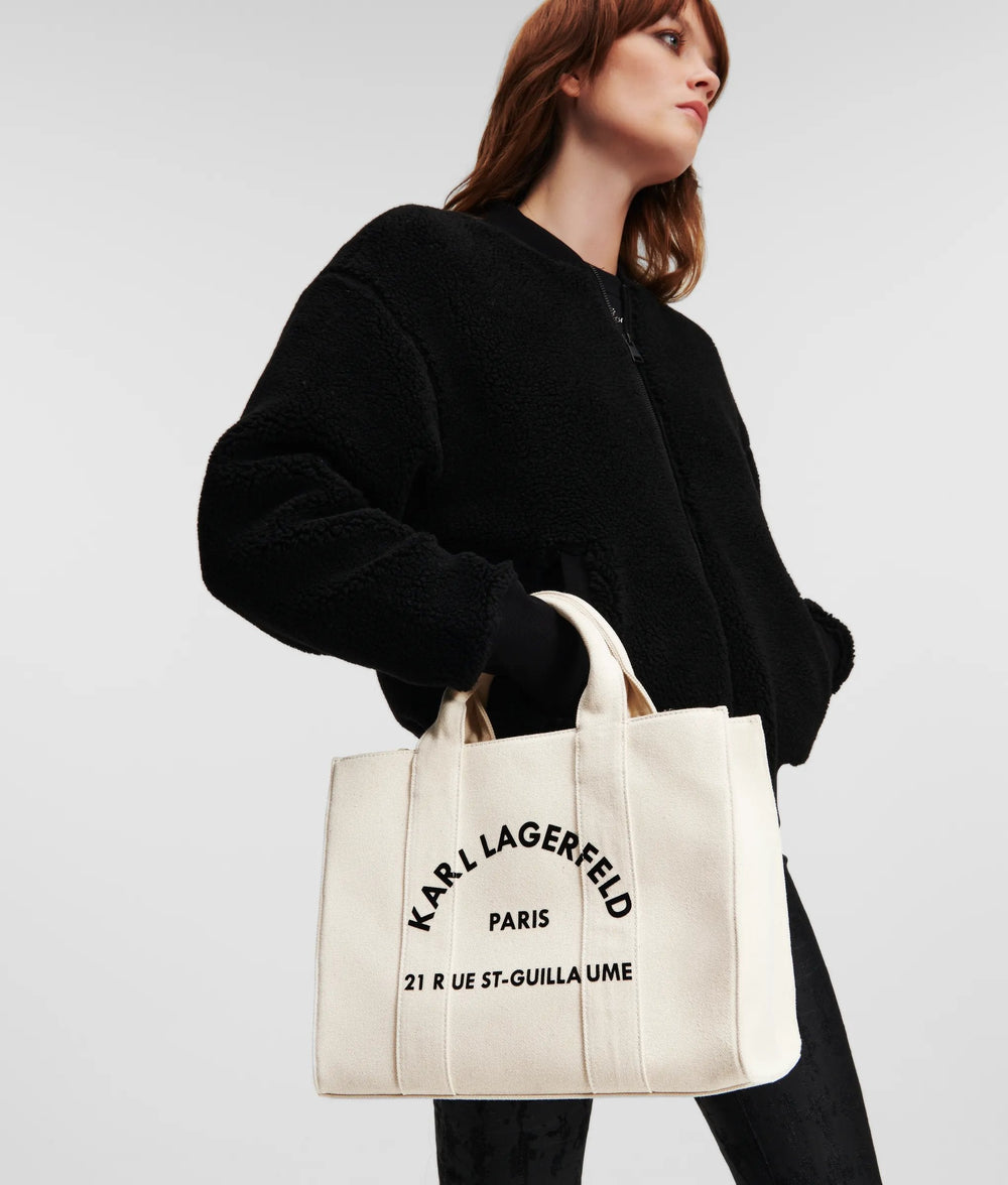 Karl Lagerfeld beige Essentials-shoppingväska, buren i handen med svart jacka och svarta byxor.