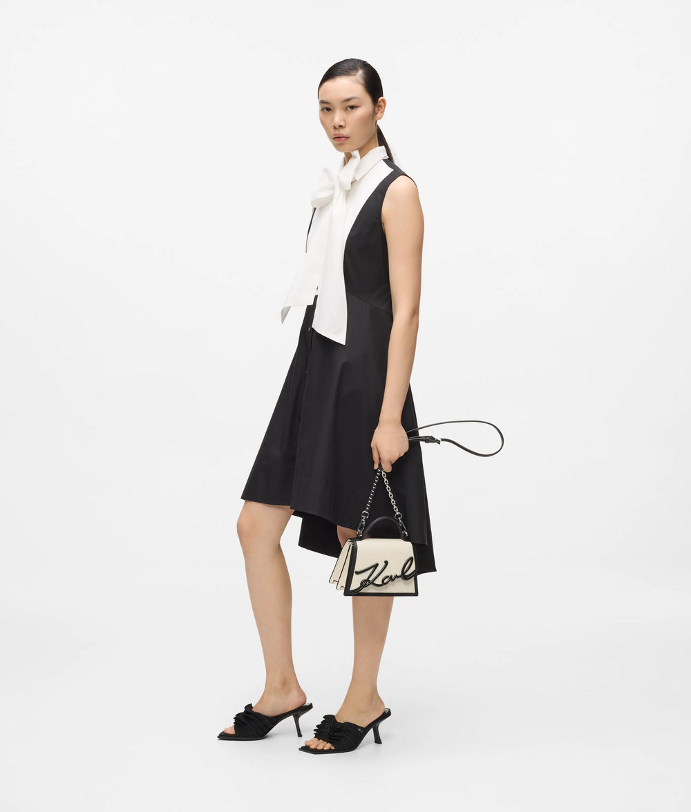 Karl Lagerfeld sac à main Essentials noir tenu à la main avec une robe noire sans manches et une blouse blanche.