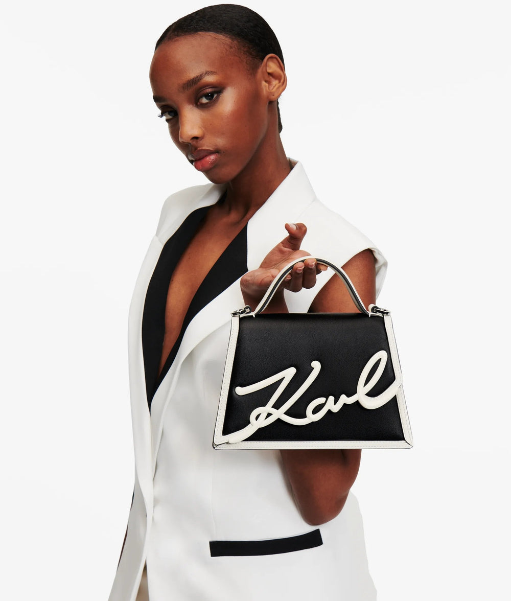 Karl Lagerfeld sac à main Essentials noir tenu à la main, avec un blazer blanc sans manches et un haut noir.