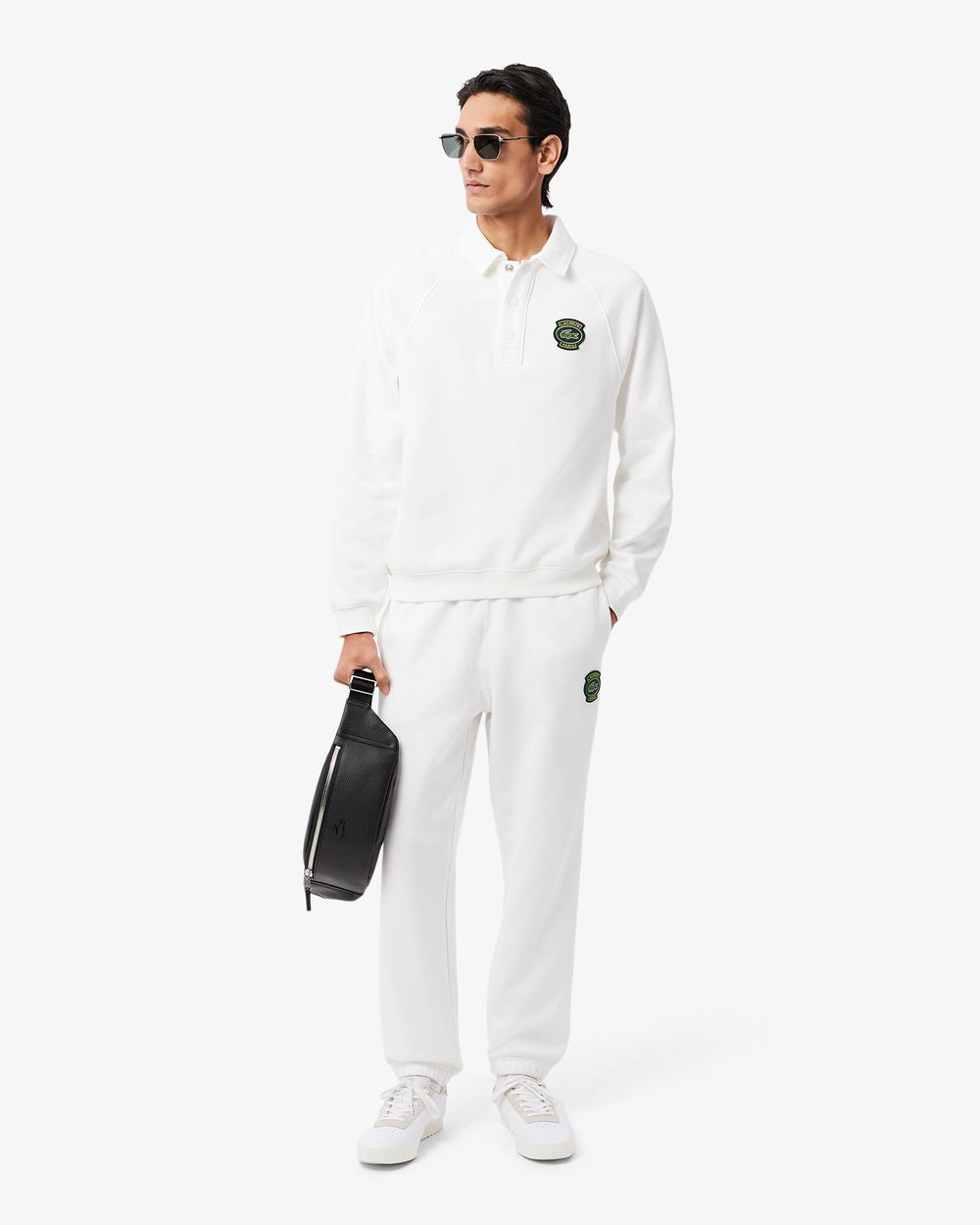 Lacoste banane Chantaco noire tenue à la main avec un ensemble blanc et des baskets blanches.