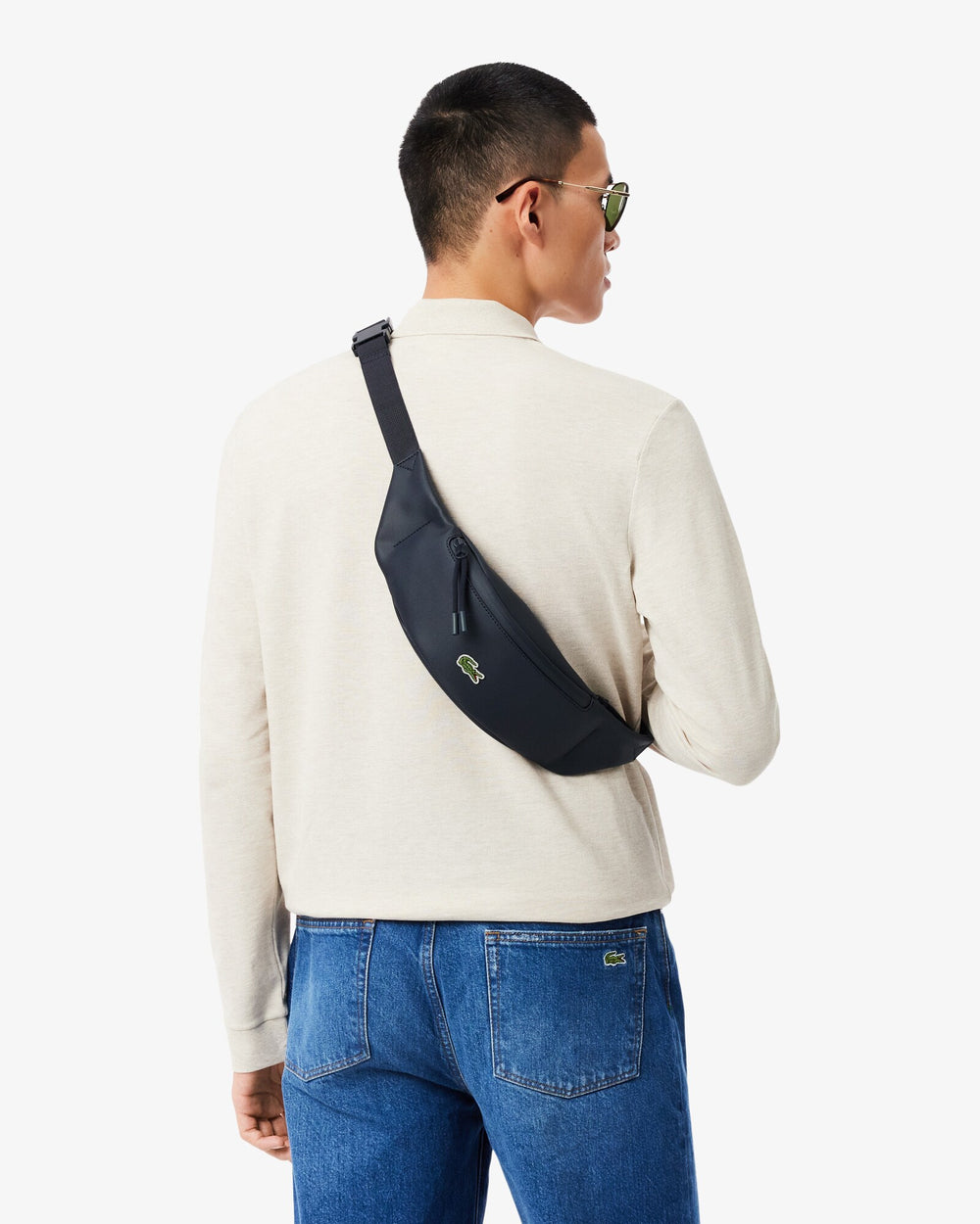 Lacoste Bauchtasche Lcst in Blau, quer über einem weißen Oberteil und blauen Jeans getragen.