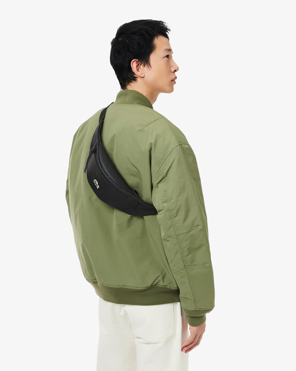 Lacoste schwarze Bauchtasche Lcst, quer über dem Rücken getragen, zu grüner Jacke und weißer Hose.