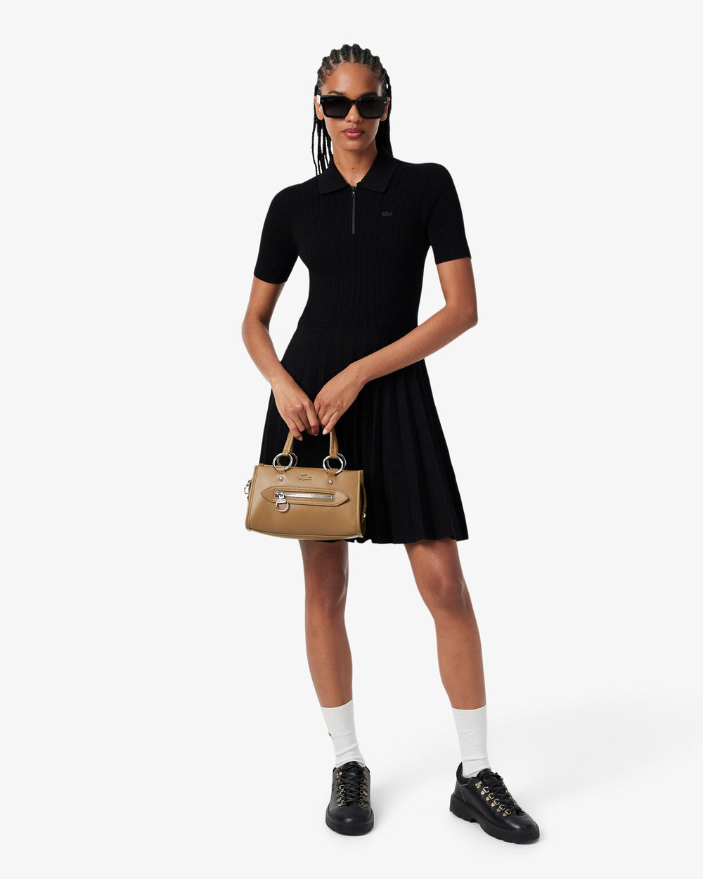 Lacoste mala Celys castanha, segurada à mão, com um vestido preto, meias brancas e sapatilhas pretas.