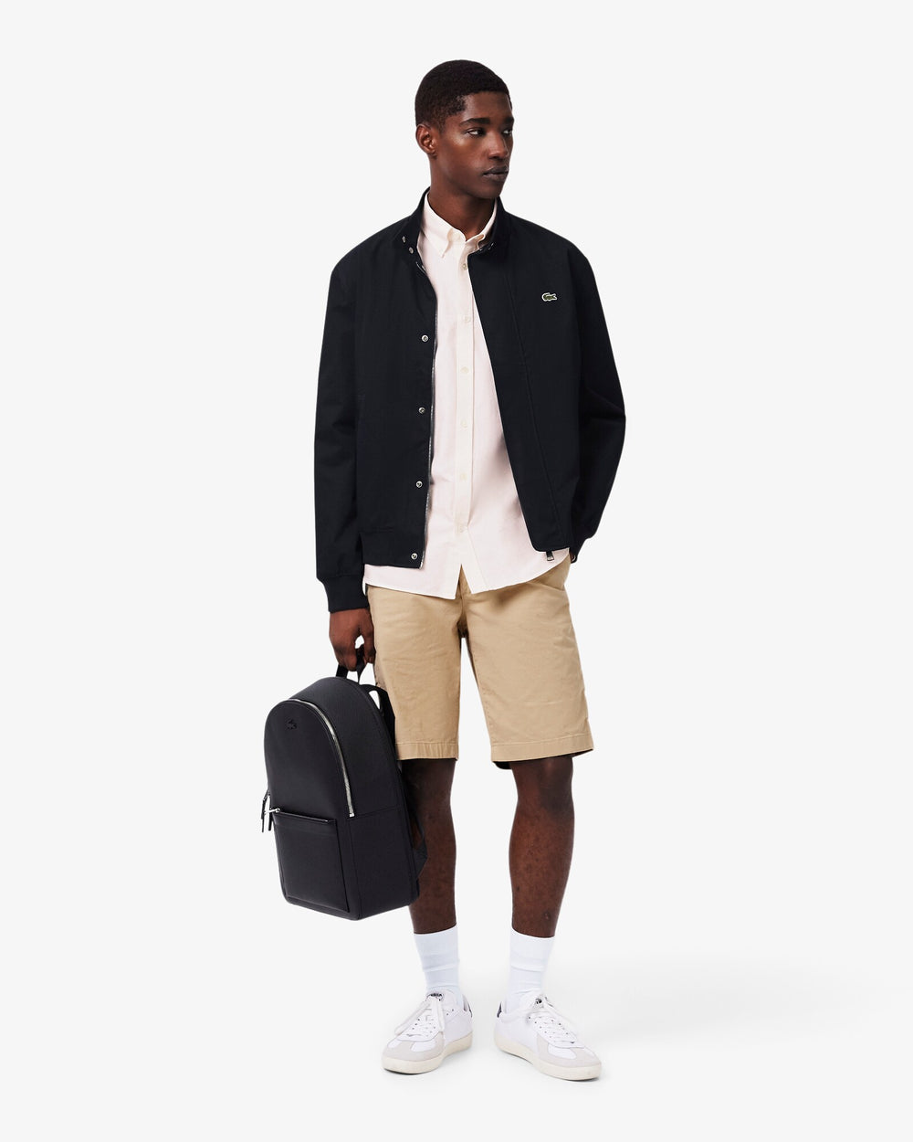 Lacoste sac à dos Chantaco noir tenu à la main, avec une veste noire, une chemise blanche et un short beige.