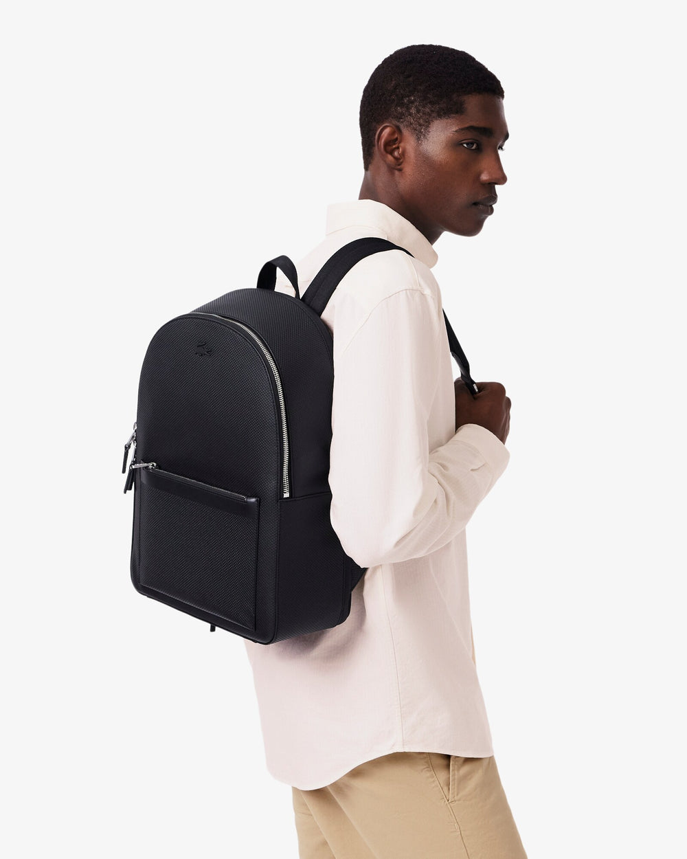 Lacoste sac à dos Chantaco noir porté sur le dos avec une chemise blanche et un pantalon beige.