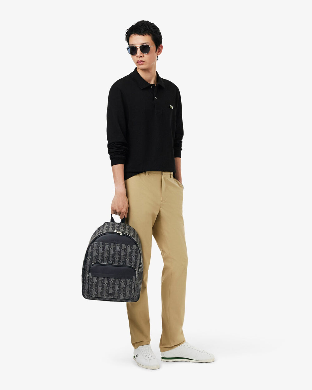 Lacoste czarny plecak Men’s Classic trzymany w ręku, z czarną koszulką polo i beżowymi spodniami.
