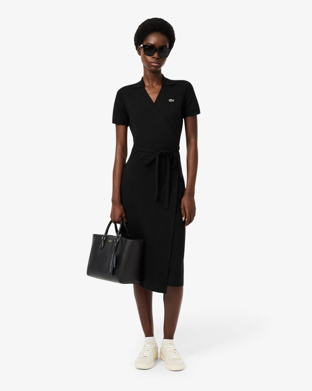 Lacoste cabas Champs Elysees noir tenu à la main, avec robe noire et baskets blanches.