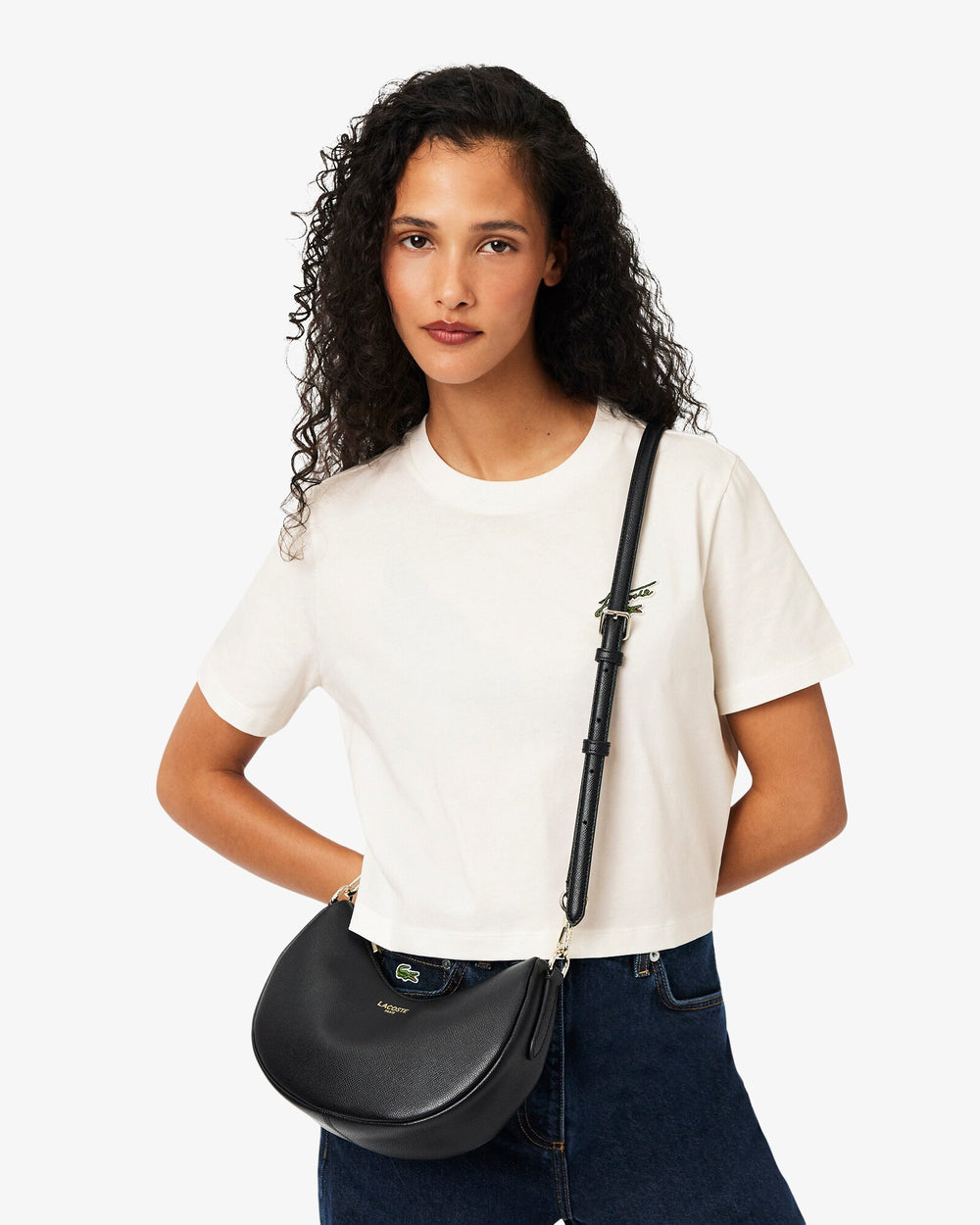 Lacoste sac demi-lune Champs Elysees noir porté en bandoulière avec un tee-shirt blanc et un jean bleu.