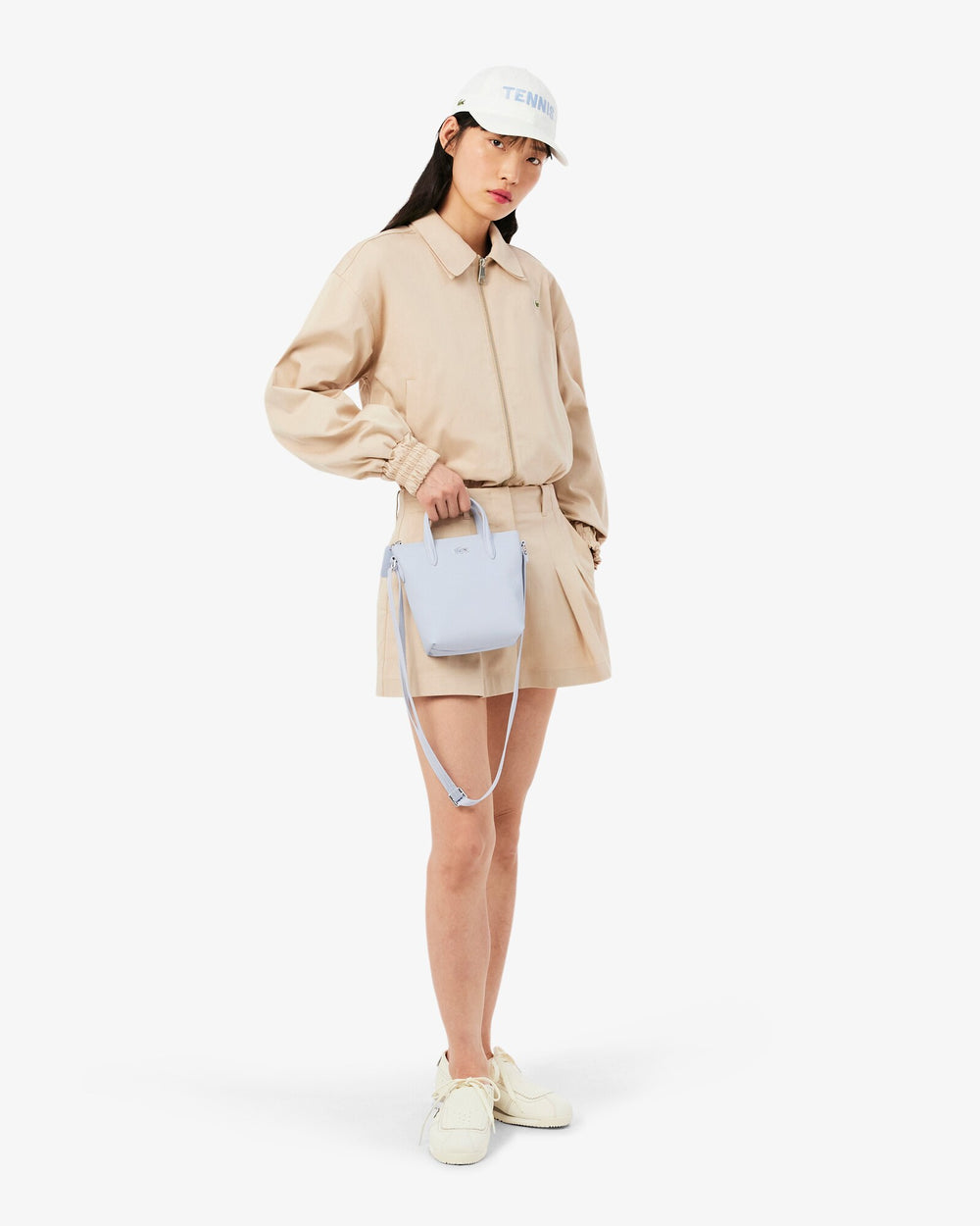 Lacoste cabas L.12.12 Concept bleu tenu à la main, avec une veste et un short beiges.