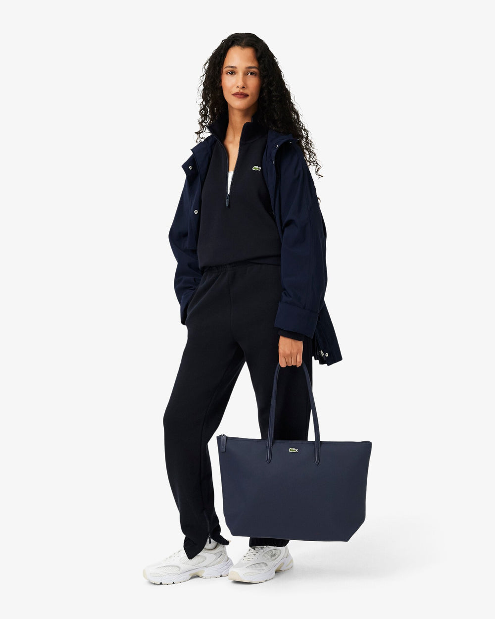 Lacoste modrá nákupná taška L.12.12 Concept držaná v ruke, s tmavomodrou bundou a čiernymi nohavicami.