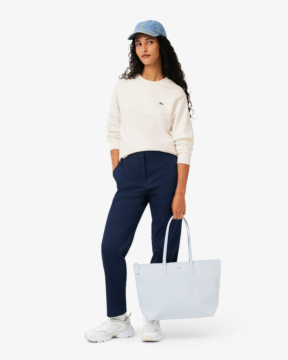Lacoste modrá nákupná taška L.12.12 Concept, nesená v ruke, s bielym svetrom a tmavomodrými nohavicami.