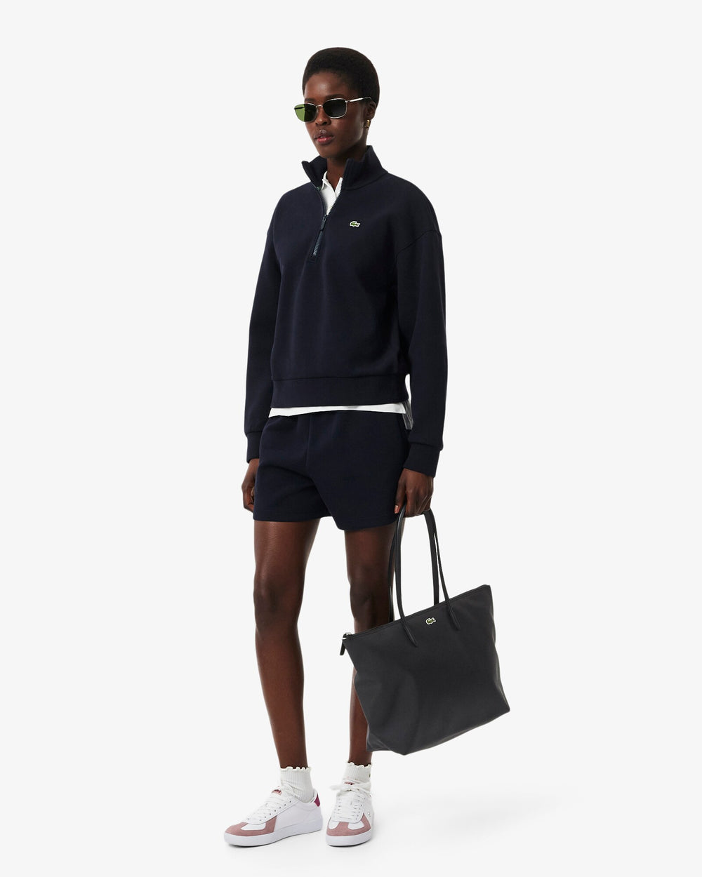 Lacoste čierna nákupná taška L.12.12 Concept, držaná v ruke k tmavomodrému vrchu, šortkám a bielym teniskám.