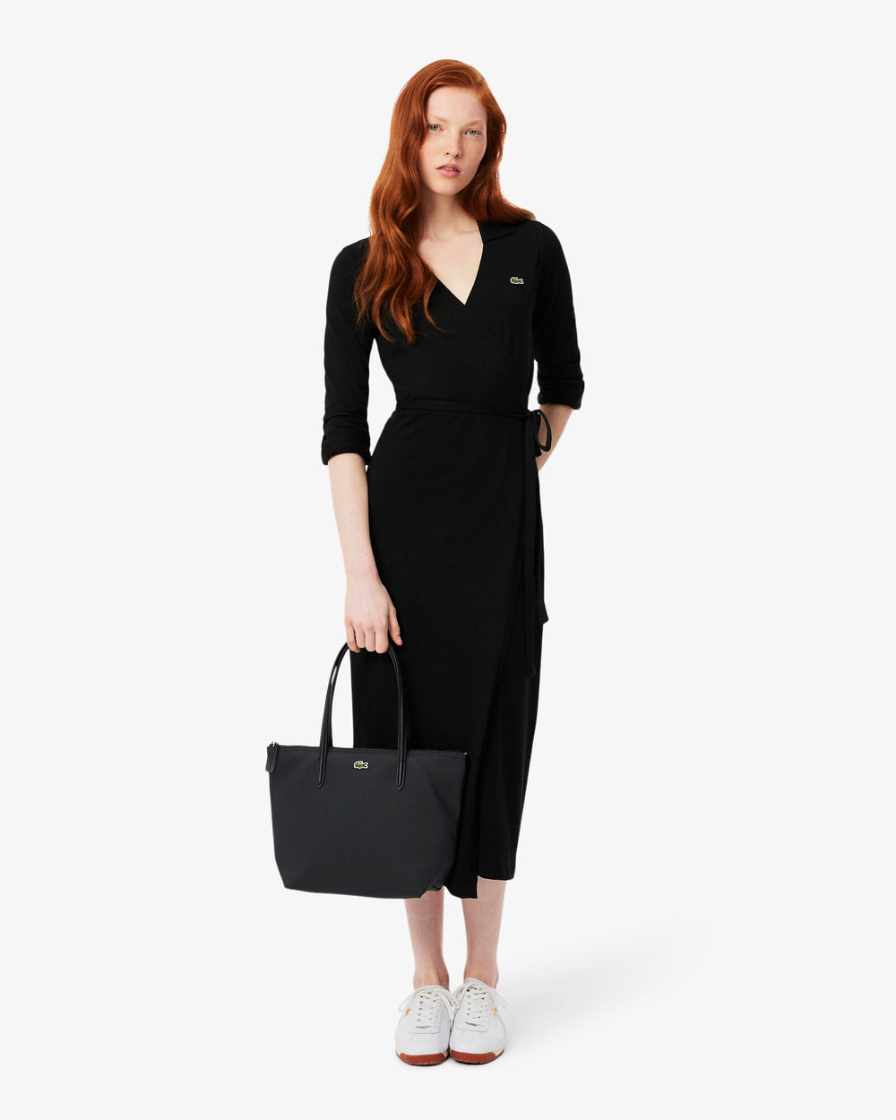 Lacoste saco de compras L.12.12 Concept preto levado à mão com um vestido preto e sapatilhas brancas.