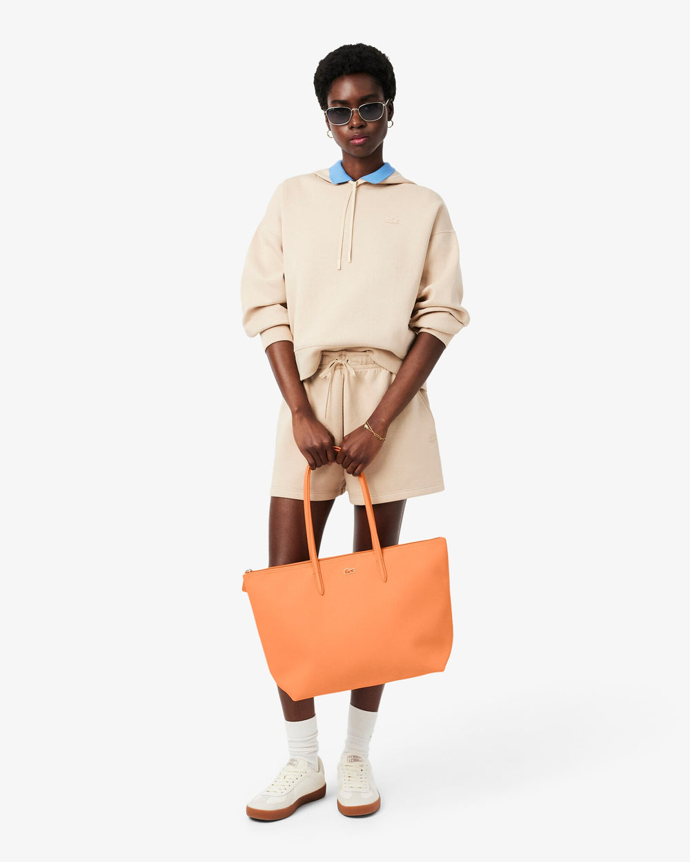 Lacoste oranžová taška L.12.12 Concept držaná v ruke pred béžovým oblečením, modrým golierom a bielymi teniskami.