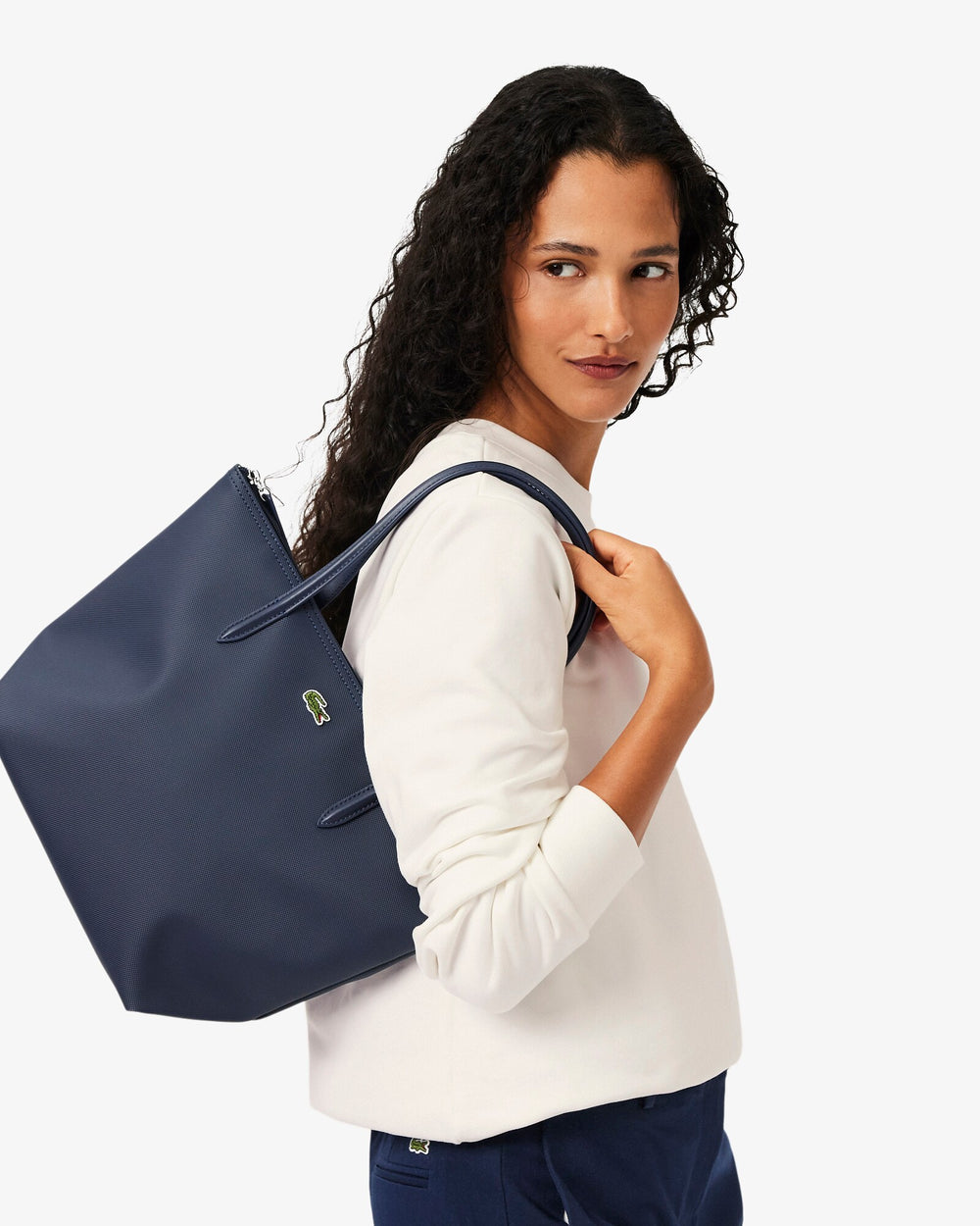 Lacoste saco L.12.12 Concept azul levado ao ombro, com uma camisola branca e umas calças azul-marinho.