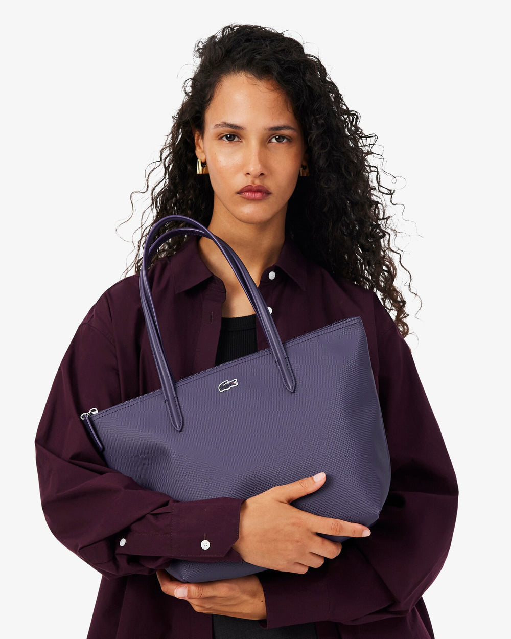 Saco de compras Lacoste L.12.12 Concept roxo, segurado à frente do corpo, com camisa cor de vinho e peça preta.