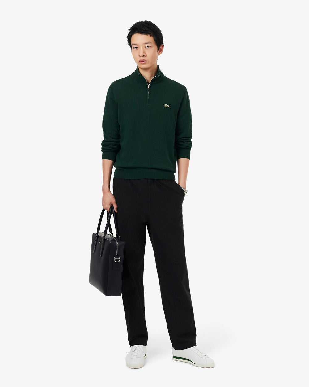 Lacoste bolso rectangular Langon negro, llevado a mano, con una prenda superior verde oscuro, pantalón negro y zapatillas blancas.