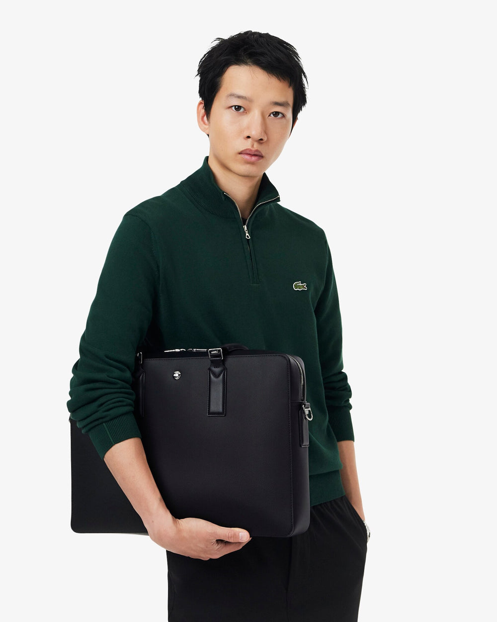 Lacoste bolso rectangular Langon negro, sujeto contra el torso con un jersey verde oscuro y un pantalón negro.