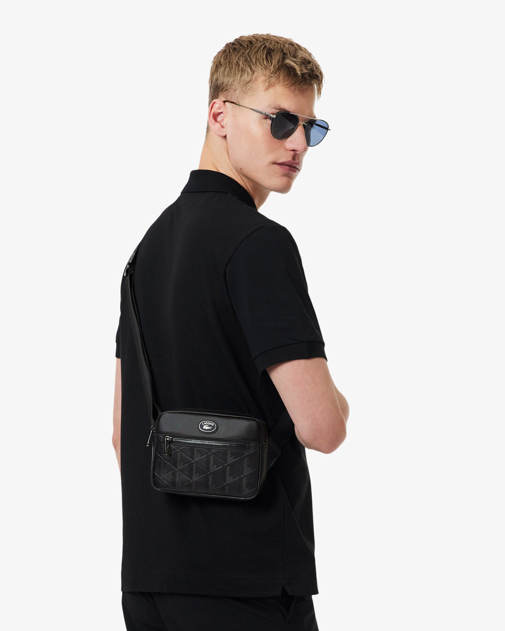 Lacoste borsa Reporter Monogramme nera, portata a tracolla con una polo nera e pantaloni neri.