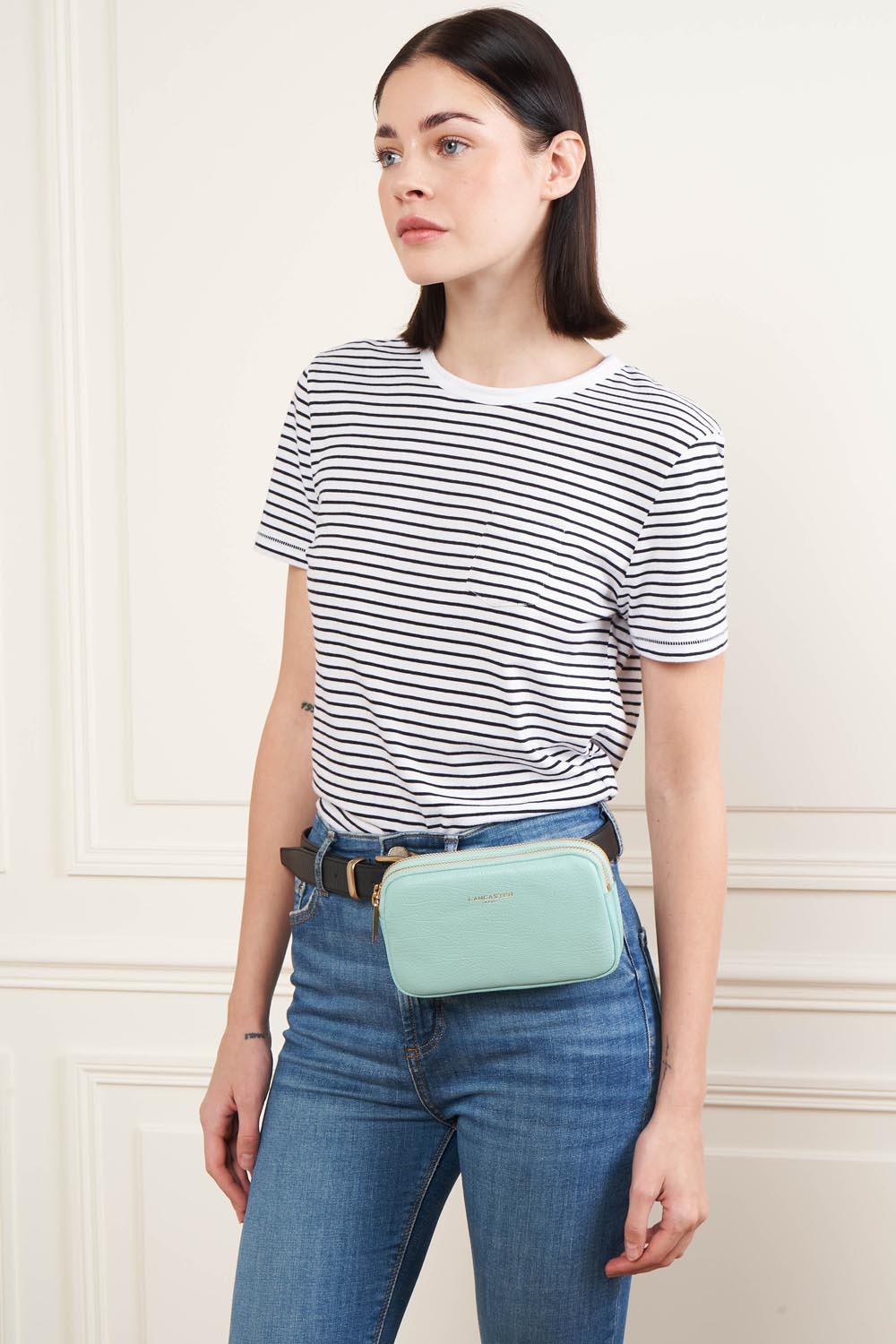 Lancaster Double pochette Zippé Dune Lagon Clair – Med en stribet T-shirt og blå jeans bæres Dune-clutchen i taljen.