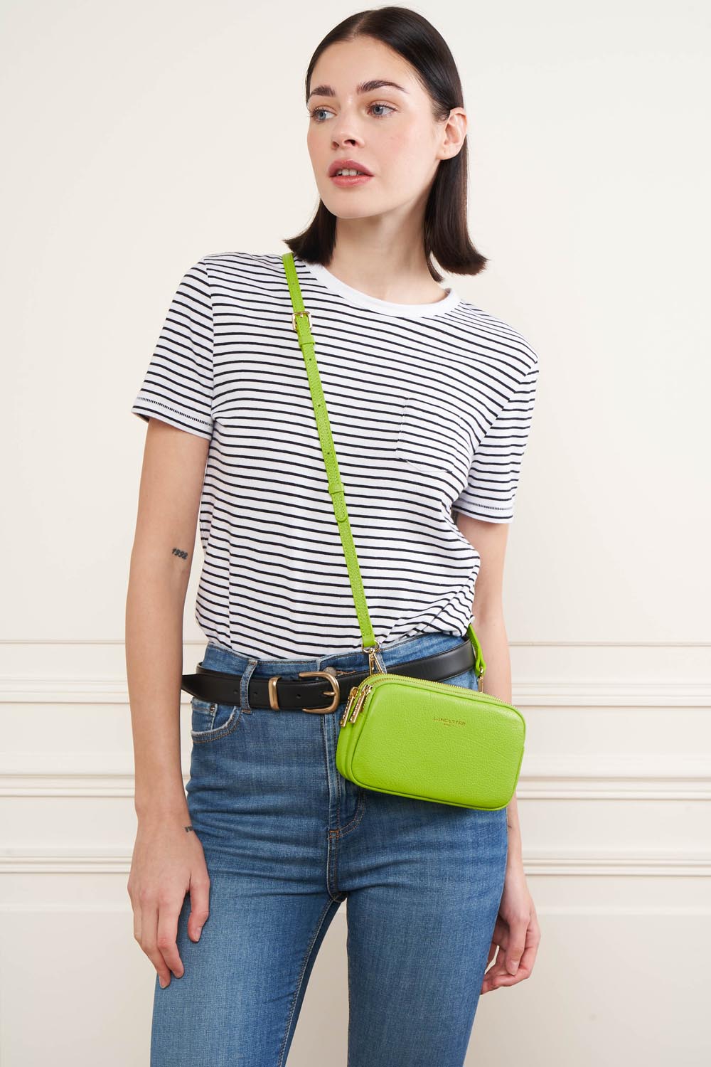 Lancaster Double pochette Zippé Dune Lagon Clair – Den grønne Dune-pung bæres på tværs af kroppen til en stribet T-shirt og blå jeans.