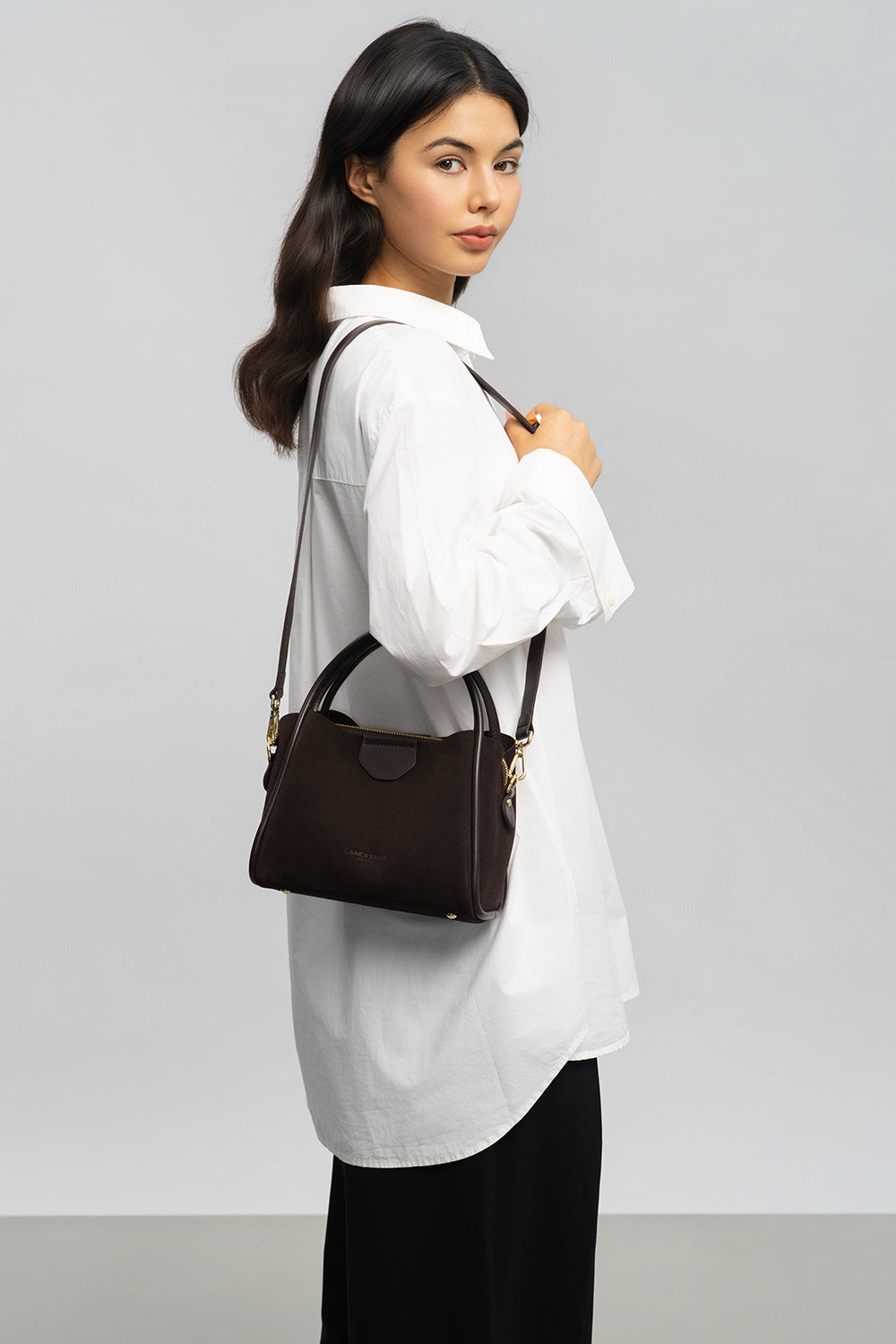 Lancaster Sac à main Aimantée S Suède Ida Taupe – Marron, le sac seau se tient à la main et complète une chemise blanche sur un pantalon noir.