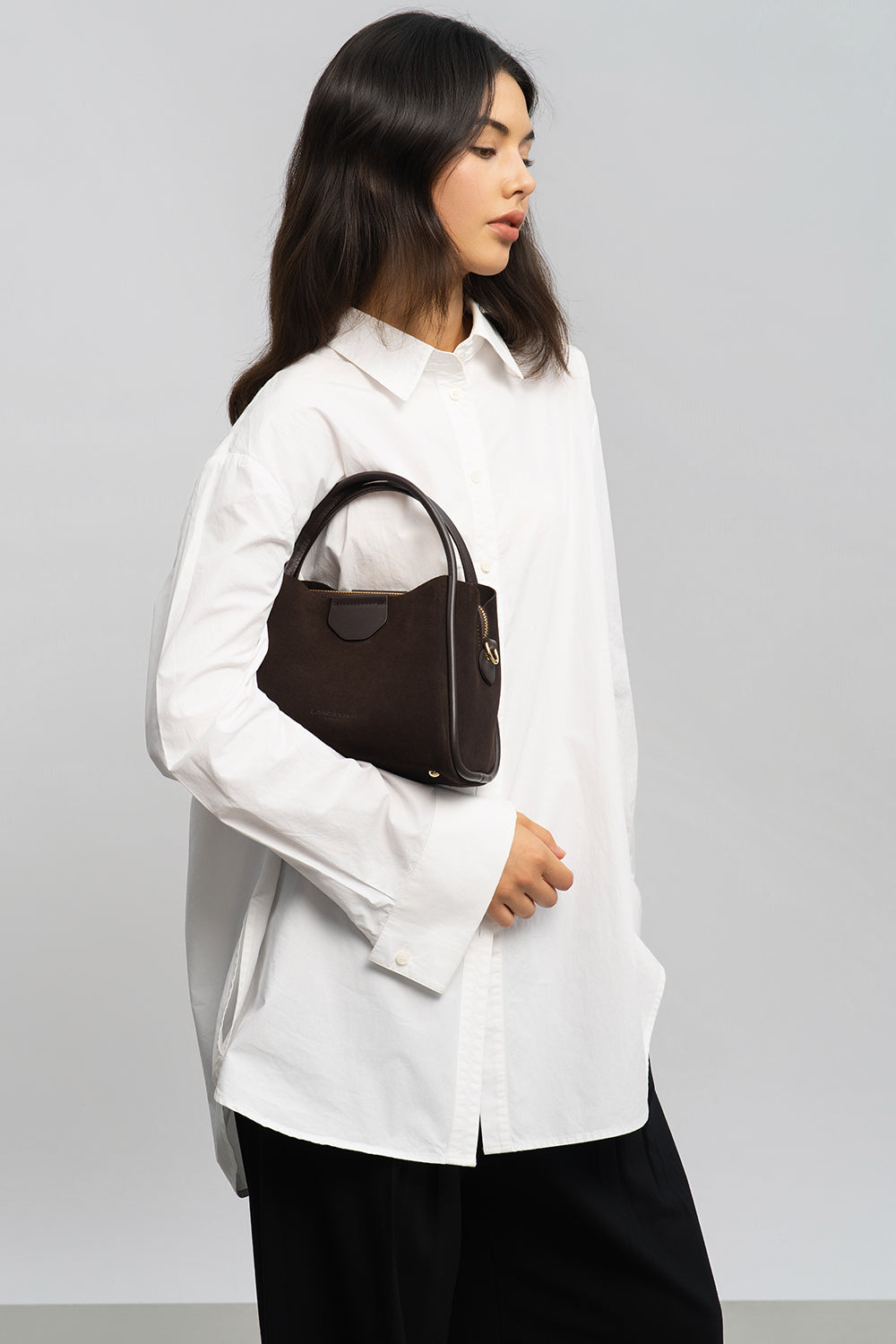 Lancaster Sac à main Aimantée S Suède Ida Taupe – À la main, le sac seau marron complète une chemise blanche et un pantalon noir.