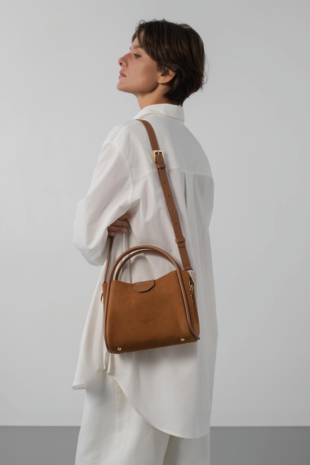 Lancaster Sac à main Aimantée S Suède Ida Taupe – Une chemise blanche et un pantalon blanc pour ce sac à main marron porté en bandoulière.