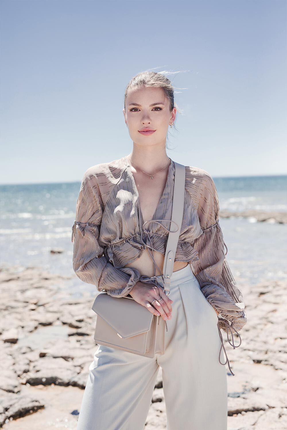 Lancaster Zoé Flap Handbag with Clasp Black – The beige Zoé handbag is worn crossbody with a taupe patterned top and white trousers.
