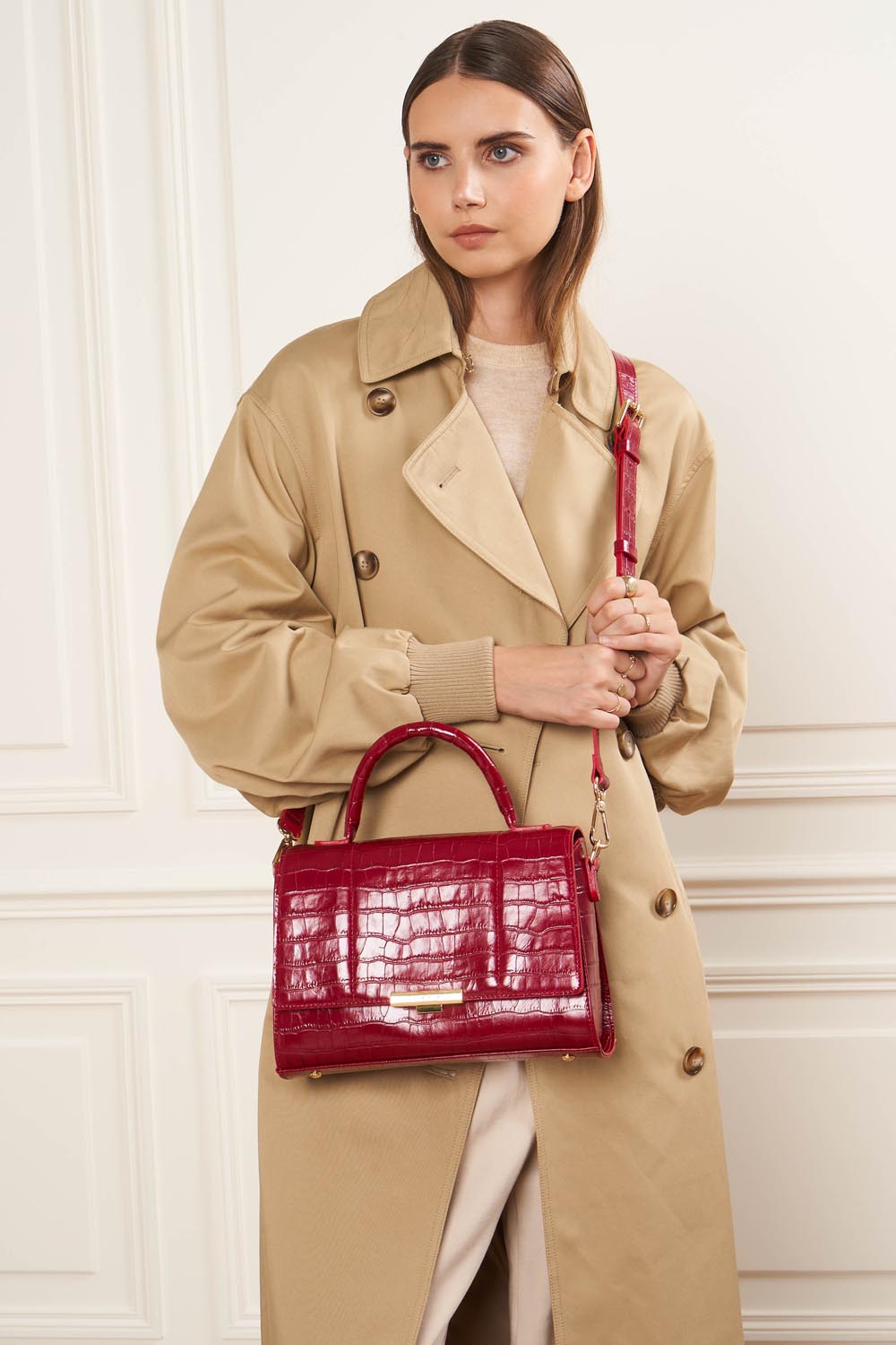 Lancaster Sac Exotic Trinity M Noir Croco – À la main, le sac à main rouge complète un trench beige et un pantalon blanc.