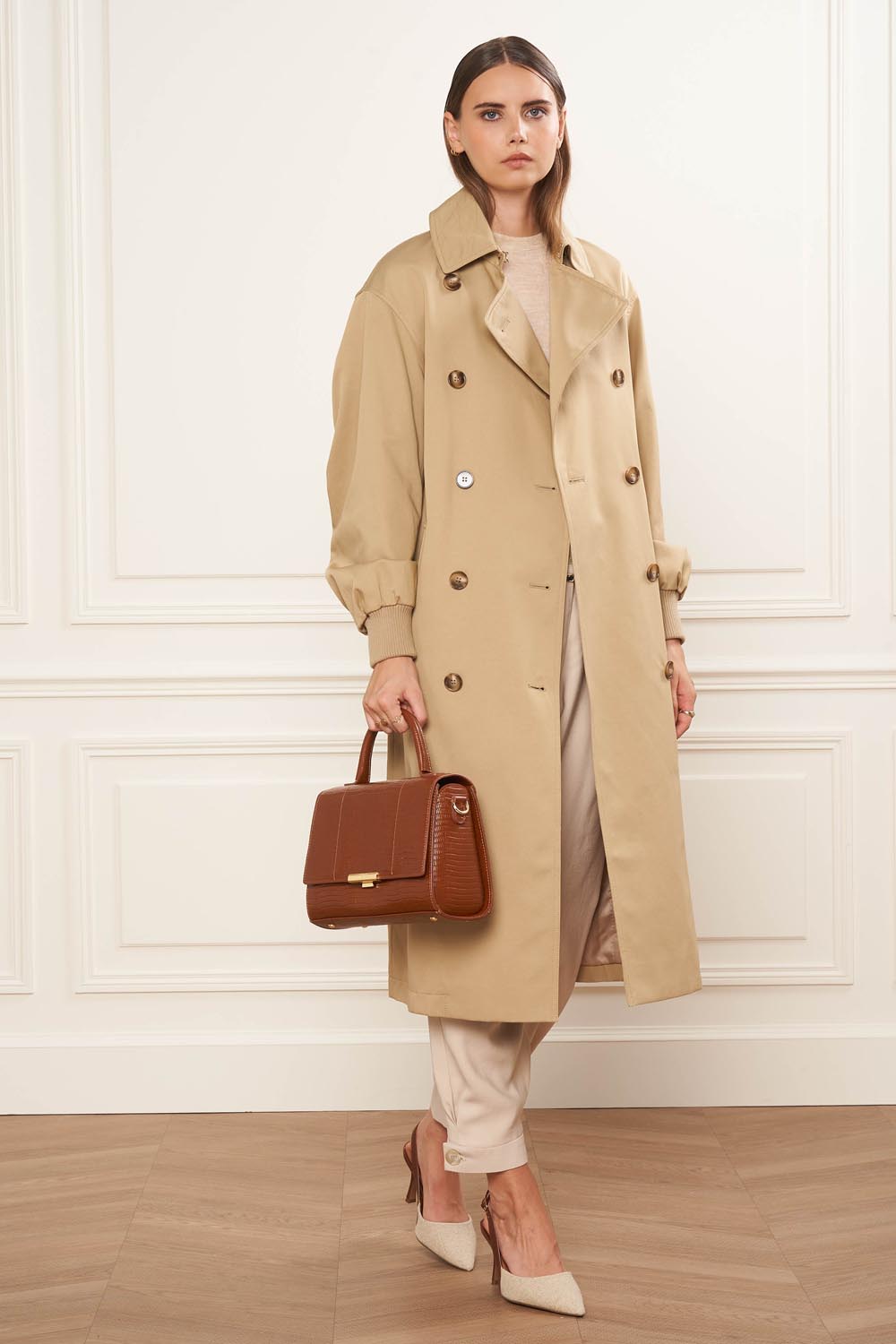 Lancaster Sac Exotic Trinity M Noir Croco – Le sac à main marron se tient à la main avec un trench beige et un pantalon clair.