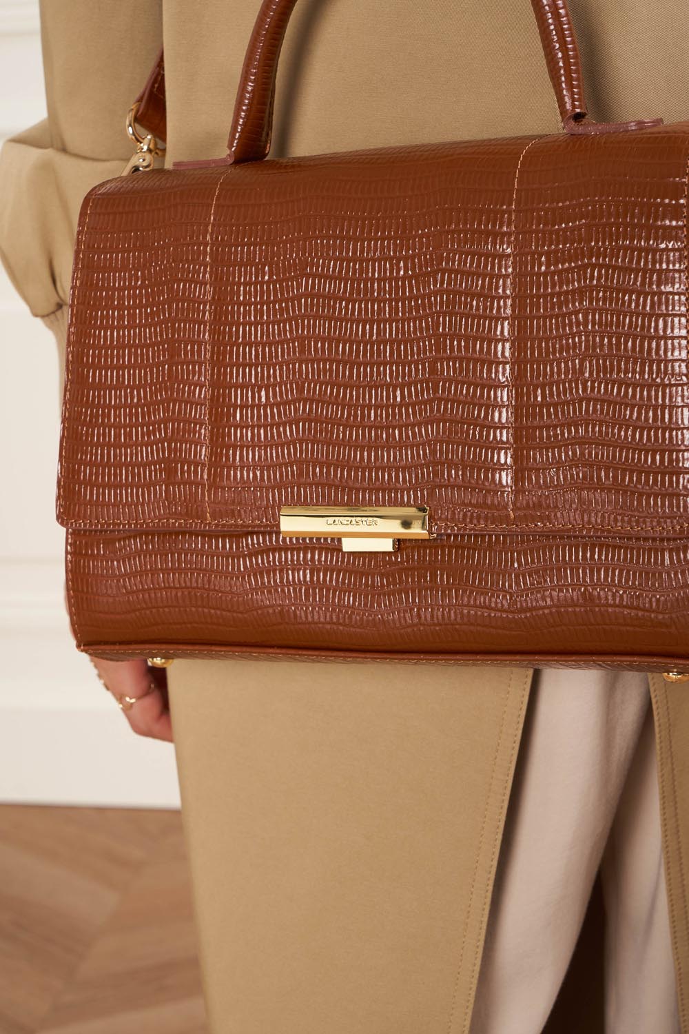 Lancaster Sac Exotic Trinity M Noir Croco – À la main, le sac à main brun s’accorde à un manteau beige et un pantalon assorti.