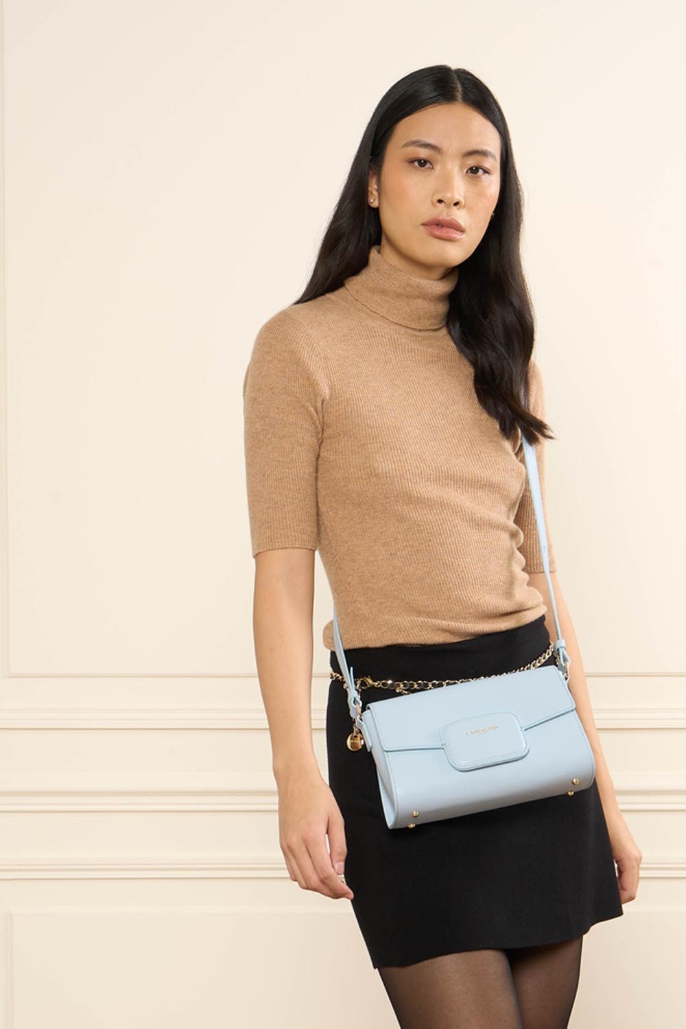 Lancaster Sac Trotteur Paris Pad Noir – Le trotteur Paris PAD se porte en bandoulière avec un haut beige et une jupe noire.