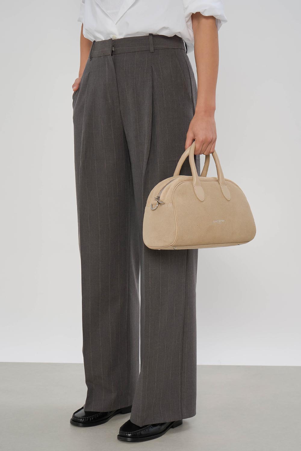 Beige Lancaster Bowling-Tasche in der Hand, dazu ein weißes Hemd und eine graue Hose.