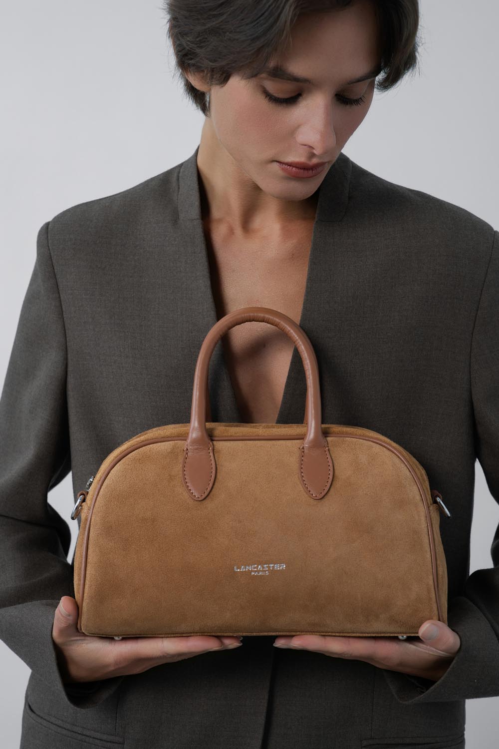 Lancaster, brown Bowling bag held in both hands in front of a grey blazer and black top.