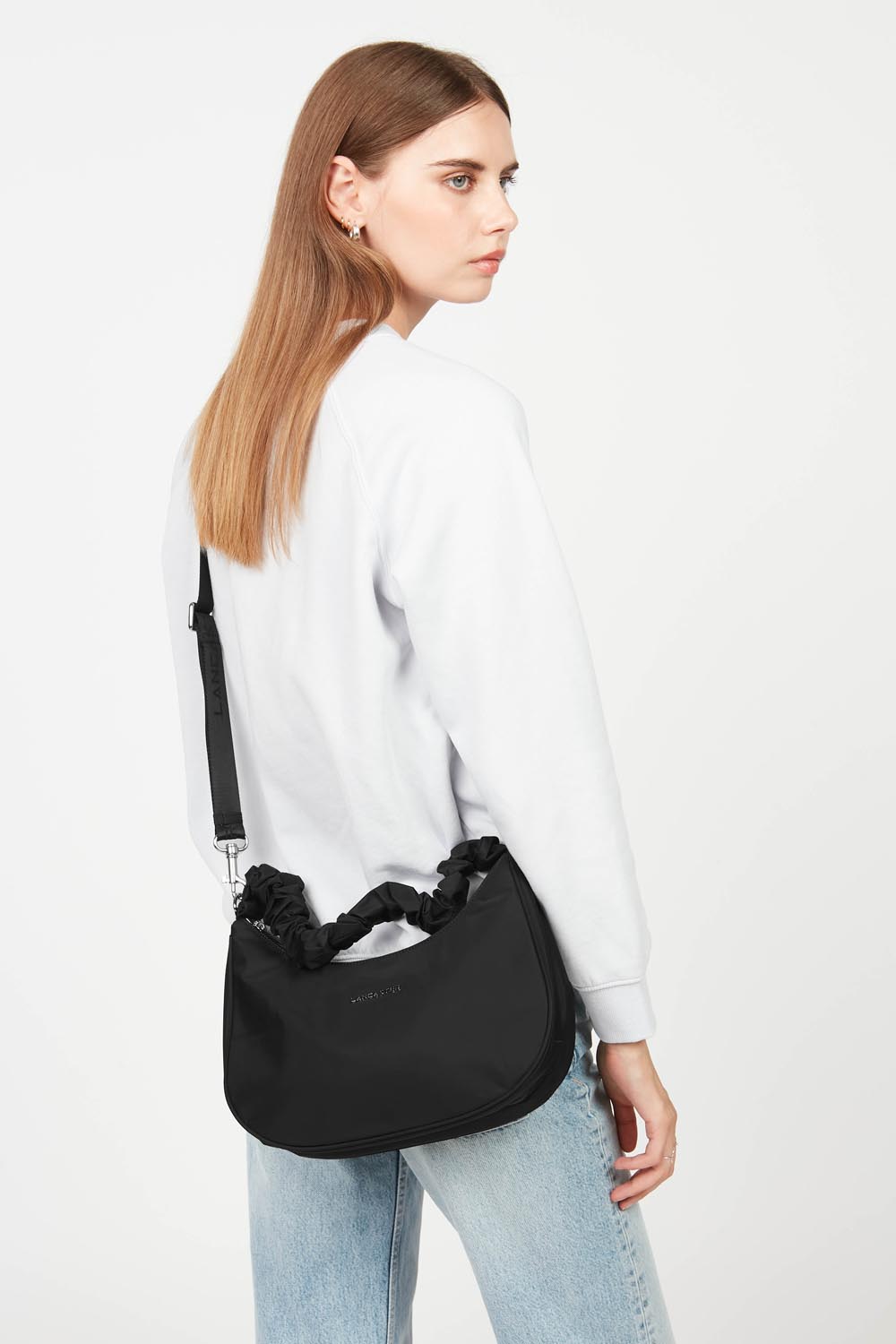 Lancaster, mala Hobo Basic Chouchou preta usada a tiracolo, com uma camisola branca e umas calças de ganga azul-claro.