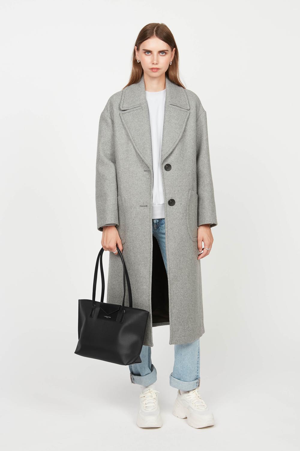 Lancaster, cabas Maya noir porté à la main avec un manteau gris et un jean bleu.